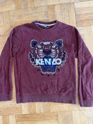 Vinröd sweatshirt från Kenzo - Cool vinröd sweatshirt från Kenzo med det ikoniska tigermotivet broderat på bröstet och texten 'KENZO PARIS'. Tröjan har rund hals, långa ärmar och ribbade muddar. Perfekt för dig som vill sticka ut med en streetwear vibe. Storlek S. Det är bara att höra av dig vid frågor!!🙋‍♂️
