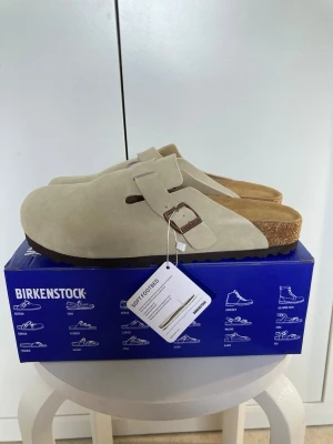 Beige Birkenstock Boston tofflor - Säljer ett par beige Birkenstock Boston tofflor i mocka med justerbart spänne i metall. Tofflorna har en klassisk korksula och mjuk fotbädd för extra komfort. Perfekta för dig som vill ha både stil och bekvämlighet i vardagen.