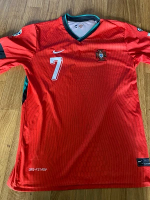 Portugal matchtröja Ronaldo #7 Nike - Säljer en röd Portugal matchtröja från Nike med Ronaldo och nummer 7 på ryggen. Tröjan har gröna detaljer vid kragen, officiellt landslagsmärke på bröstet och är tillverkad i Dri-FIT material som andas. Perfekt för dig som älskar fotboll och vill stötta Portugal.