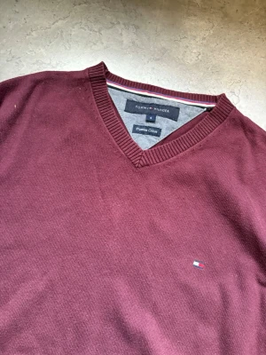 Vinröd stickad tröja Tommy Hilfiger - Vinröd stickad tröja från Tommy Hilfiger med V-ringning och diskret logga broderad på bröstet. Tröjan har långa ärmar, ribbade muddar och är gjord i mjuk bomull. Perfekt för lager på lager och enkel att matcha.