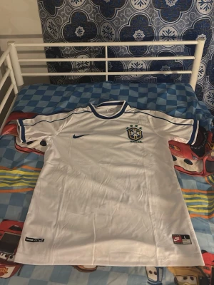 Vit Brasilien Nike fotbollströja - Snygg vit fotbollströja från Nike med Brasilien-emblem på bröstet och blå detaljer vid krage och ärmslut. Tröjan har korta ärmar och är tillverkad i ett lätt och ventilerande material, perfekt för träning eller match.