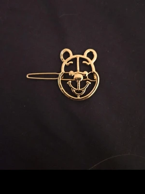 Guldig hårklämma med nalle puh - Söt hårklämma i metall med ett guldigt björnmotiv. Klämman har en öppen design som formar ett leende björnhuvud och ger en lekfull touch till din frisyr. Perfekt accessoar för att piffa upp håret med en unik detalj. Aldrig använd.