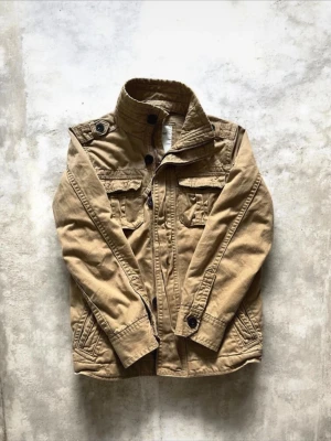 Beige utilityjacka Abercrombie & Fitch - Snygg beige utilityjacka från Abercrombie & Fitch med flera fickor, axelklaffar och ståkrage. Jackan har knappar och dragkedja framtill samt detaljerade sömmar. Perfekt för dig som gillar en avslappnad men ändå stilren look.