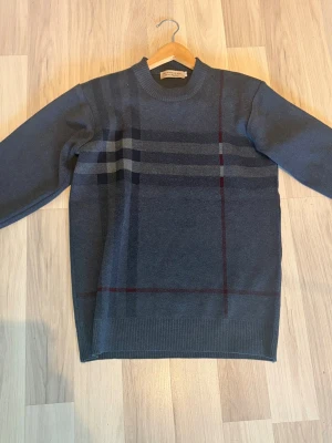 Mörkgrå stickad burberry tröja - Mörkblå stickad tröja med diskret rutmönster i grått och vinrött. Tröjan har rund halsringning, långa ärmar och ribbade muddar vid ärmslut och nederkant. Perfekt för dig som gillar klassisk och stilren design.