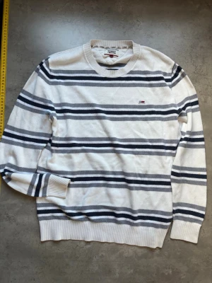 Randig tröja från Tommy Jeans - Vit tröja från Tommy Jeans med horisontella ränder i mörkblått och grått. Tröjan har rund halsringning, ribbade muddar och en liten broderad logga på bröstet. Perfekt för en avslappnad och stilren look. Mindre fläck på ena armen 