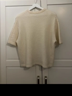 Krämvit stickad t-shirt - En krämvit stickad t-shirt med korta ärmar och rund hals. Tröjan har en avslappnad passform och ribbade kanter vid ärmslut och nederkant. Perfekt för en stilren och enkel look.
