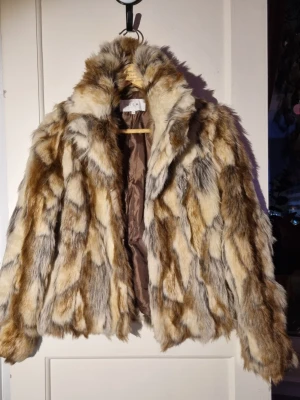 Beige och brun fuskpälsjacka - Säljer en fluffig fuskpälsjacka i beige, brunt och grått med coolt mönster. Jackan har hög krage, långa ärmar och är fodrad på insidan för extra värme. Perfekt för dig som vill sticka ut och hålla dig varm under vintern.