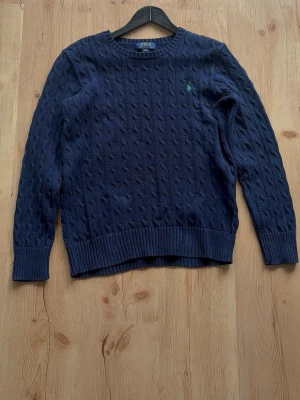 Stickad Polo Ralph Lauren  - Kabelstickad mörkblå tröja från Polo Ralph Lauren med rund halsringning och ribbade muddar. Klassisk grön broderad logga på bröstet. Tillverkad i mjukt material som passar perfekt till höst och vinter. Mer M i storlek men skulle nog passa någon i L