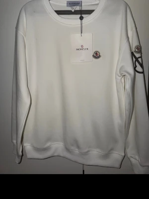 Moncler vit sweatshirt  - Säljer en stilren vit sweatshirt från Moncler med rund halsringning och broderad Moncler-logga på bröstet. Tröjan har ribbade muddar vid ärmslut och nederkant samt en diskret logga på ärmen. Perfekt för en clean och trendig look.
