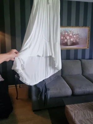 Vit drömlik långklänning med volang - Luftig vit långklänning med volangdetaljer och asymmetrisk nederkant. Klänningen har ett bohemiskt uttryck och är tillverkad i ett lätt tyg som ger ett fint fall. Perfekt för sommarens alla tillfällen.