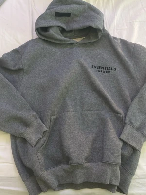 Grå Essentials Fear of God hoodie - Säljer en grå hoodie från Essentials Fear of God med svart logga på bröstet. Hoodien har stor huva, magficka och ribbade muddar vid ärmar och midja. Tillverkad i mjukt och tjockt material som känns riktigt skönt mot huden.