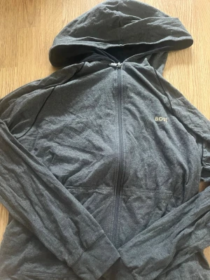 Mörkgrå hoodie med dragkedja från BOSS - Snygg mörkgrå hoodie från BOSS med dragkedja framtill och guldfärgad logga på bröstet. Tillverkad i mjukt bomullsliknande material och har praktisk huva samt fickor framtill. Perfekt för en avslappnad och stilren look.