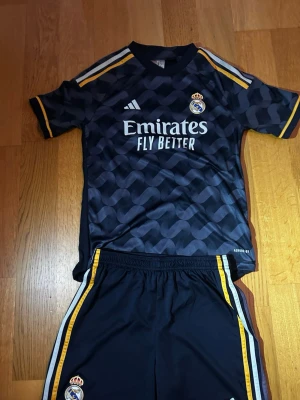 Real Madrid svart matchställ Adidas - Säljer ett svart Real Madrid matchställ från Adidas med gula och vita detaljer. Tröjan har mönstrad framsida, klubbmärke och sponsortryck. Shortsen har matchande ränder och klubbmärke. Perfekt för fotbollsträning eller match.