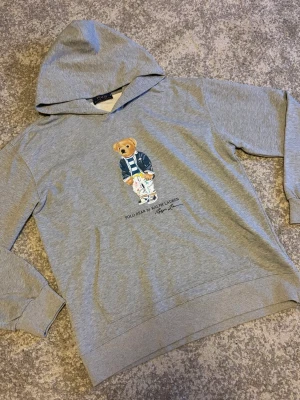 Grå hoodie Polo Bear Ralph Lauren - Säljer en grå hoodie från Polo Ralph Lauren med den ikoniska Polo Bear-trycket på bröstet. Hoodien har huva, ribbade muddar och är tillverkad i mjuk bomull. Perfekt för en avslappnad och cool stil.