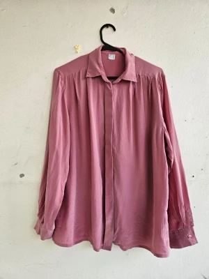 Silkesskjorta - Jättefin rosa sidenblus i tunt och härligt material. Fint skick men har några tecken på användning, den är lagad på insidan kring handlederna (bild 4), men det syns ej på utsidan. Vänster ärm är även lagad lite på utsidan ovanför knappen, vilket syns lite grann (bild 6). Den är lätt missfärgad längst nere på framsidan (bild 5) och har en jätteliten mörkrosa fläck på framsidan till vänster om knapparna (bild 7).