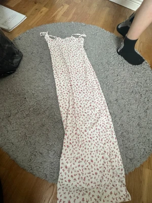 Vit långklänning med blommor - Supersöt långklänning från FB Sister i vit med små rosa blommor och gröna blad. Klänningen har smala axelband med knyt och en liten rosett framtill. Perfekt för sommarens alla tillfällen!