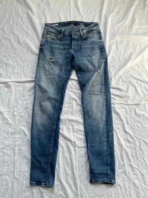  jeans från Jack & Jones -  jeans från Jack & Jones i ljusblå tvätt med slitningar och distressed detaljer. Modellen heter Glenn och har klassisk femficksdesign, dragkedjegylf och kontrastsömmar. Perfekta för en avslappnad och trendig look.