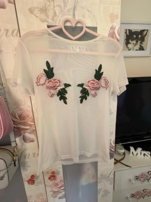 Vit mesh topp med blommor från ISTAY - Säljer en transparent vit mesh topp från ISTAY med broderade rosa blommor och gröna blad framtill. Toppen är kortärmad och har en loose passform, perfekt för att styla lager på lager. Superfin detalj med blommorna som ger en unik look.