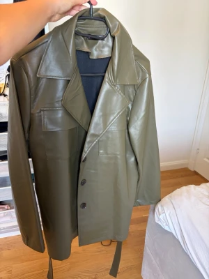 Olivgrön lång skinnjacka med bälte - Snygg olivgrön skinnjacka med lång passform och bälte i midjan. Jackan har breda slag, knäppning framtill med svarta knappar och två bröstfickor. Perfekt för dig som vill sticka ut med en trendig look.