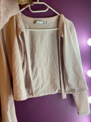 Ljusrosa kofta med knytband - Ljusrosa kofta med lång ärm och knytband i halsen som kan formas till en rosett. Koftan har en rak och avslappnad passform och är gjord i ett mjukt, lätt texturerat material. Perfekt att bära över en topp för en söt och enkel stil.