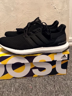 Svarta Adidas Ultra Boost sneakers - Svarta Adidas Ultra Boost sneakers med stickad ovandel och vita Boost-sulor. Skorna har svarta plastdetaljer på sidorna och hälpartiet samt klassisk snörning. Perfekta för dig som gillar sportig och stilren look.