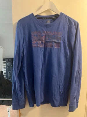 Blå långärmad topp med tryck - Säljer en blå långärmad topp med tryck framtill där det står 'New Zealand Auckland 25' och har grafiska detaljer. Toppen har rund halsringning och är gjord i ett mjukt material som känns skönt mot huden. Perfekt för en avslappnad stil.