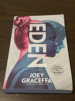 Children of Eden av Joey Graceffa - En fängslande dystopisk roman av Joey Graceffa om Rowan, en andra född i en värld där befolkning är strikt kontrollerad. Boken har ett färgstarkt omslag med porträtt i blått och rosa. Perfekt för dig som gillar spännande framtidsskildringar och äventyr.