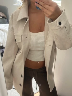 Beige kappa - Stilren beige kappa med klassisk krage. Modellen är rak och har en bröstficka med lock. Materialet ser ut att vara mjukt och lite tjockare, perfekt för lager på lager. Passar till många olika outfits.