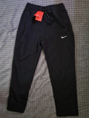 Svarta mjukisbyxor från Nike - Svarta mjukisbyxor från Nike med klassisk vit logga på vänster ben. Byxorna har resår i midjan med dragsko och raka ben. Tillverkade i mjukt och bekvämt material, perfekta för chill eller träning. Snygg och enkel design som funkar till det mesta.
