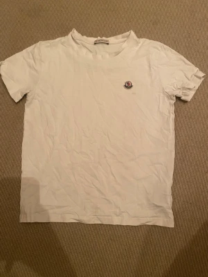 Vit t-shirt från Moncler - Säljer en klassisk vit t-shirt från Moncler med liten logga på bröstet. T-shirten har rund hals och korta ärmar, perfekt för en clean och stilren look. Tillverkad i mjuk bomull som känns skön mot huden. NFC har slutat funka men qr koden funkar. Tveka inte att höra av dig.        Mvh Sam