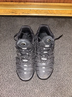Svarta Nike tns Utilitys - Snygga svarta Nike sneakers med reflekterande detaljer och ett unikt mönster över hela skon. Skorna har en robust sula och klassisk snörning. Perfekta för dig som vill ha en sportig och modern look.