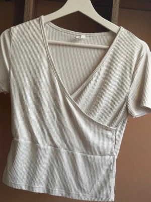 Vit omlottopp med smala ränder - Snygg vit omlottopp med smala vertikala ränder och korta ärmar. Toppen har en v-ringad omlottdesign och är tillverkad i ett ribbat material som ger en skön och stretchig känsla. Perfekt till jeans eller kjol för en fräsch look.