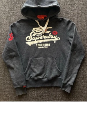Mörkgrå Superdry hoodie med tryck - Mörkgrå hoodie från Superdry med stor vit logga och text på bröstet samt röd detalj på ärmen. Klassisk känguruficka framtill, ribbade muddar och vit dragsko i huvan. Mjuk bomullsblandning och avslappnad passform.