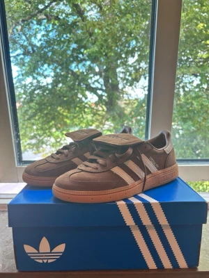 Bruna Adidas Handball Spezial Sneakers - Säljer ett par bruna Adidas sneakers med klassiska vita ränder på sidorna och gummisula. Skorna har en låg profil och är tillverkade i mocka och textil. Tungan har Adidas-logga och modellen har retrovibbar. Perfekta för dig som gillar streetwear.
