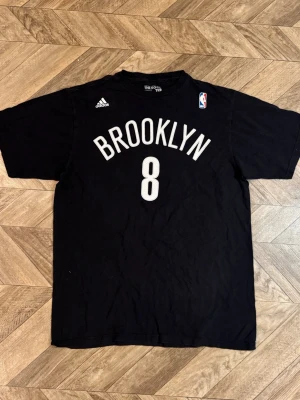 Svart Brooklyn Nets t-shirt - Säljer en svart Brooklyn Nets t-shirt från Adidas med tryck i vitt och NBA-logga på bröstet. Klassisk rund hals och korta ärmar. Perfekt för dig som gillar basket och vill ha en sportig stil.