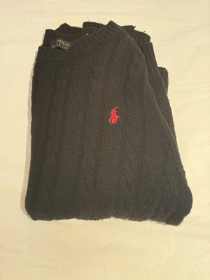 Svart kabelstickad tröja Polo Ralph Lauren - Kabelstickad svart tröja från Polo Ralph Lauren med rund halsringning och den klassiska röda logotypen broderad på bröstet. Tröjan har ribbade muddar vid ärmslut och nederkant. Perfekt för dig som gillar stilrena och tidlösa plagg.