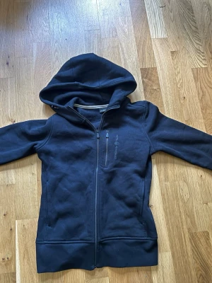 Sail racing - Snygg svart hoodie från Sail Racing med dragkedja framtill och en extra bröstficka med zip. Hoodien har en stor huva och diskret logga på bröstet. Tillverkad i mjukt material som känns skönt mot huden. Perfekt för en avslappnad och sportig stil.