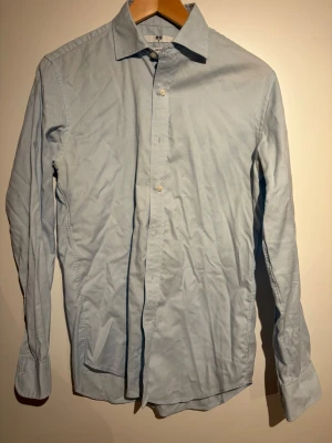 Ljusblå slim fit skjorta från Uniqlo - Säljer en ljusblå skjorta från Uniqlo i slim fit-modell. Skjortan har klassisk krage, långa ärmar och knäppning framtill. Tillverkad i bomull för en skön känsla och stilren look. Perfekt till jeans eller chinos!