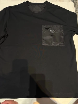 Svart Prada t-shirt med ficka och dragkedja  - Snygg svart t-shirt med rund hals och en unik bröstficka i nylon med dragkedja och en liten metallogo. T-shirten har korta ärmar och en clean, modern look som passar perfekt till jeans eller shorts.