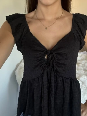 Svart topp med knyt och volang - Snygg svart topp med v-ringning, knyt framtill och volangkant nedtill. Toppen har korta ärmar med lätt volang och är tillverkad i ett mjukt, skrynkligt material som ger en avslappnad look. Perfekt att styla med jeans eller kjol.