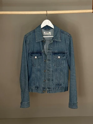 Blå jeansjacka från Acne Studios - Klassisk blå jeansjacka från Acne Studios med raka linjer, två bröstfickor med lock och knappar framtill. Jackan har krage, långa ärmar och är tillverkad i bomull. Smal passform och ganska kort i midjan.