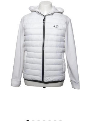 Vit pufferjacka från Hollister - Snygg vit pufferjacka från Hollister med huva och dragkedja framtill. Jackan har quiltad front och rygg, samt mjuka ärmar med trycket 'Hollister' på ena ärmen och huvan. Perfekt för kalla dagar och riktigt trendig look.