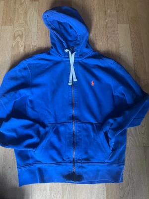 Blå zip hoodie med huva - Snygg blå hoodie med dragkedja och vit dragsko i huvan. Klassisk känguruficka framtill och en liten röd logga broderad på bröstet. Perfekt för en avslappnad och sportig stil. Mjuk och skön bomullskänsla.
