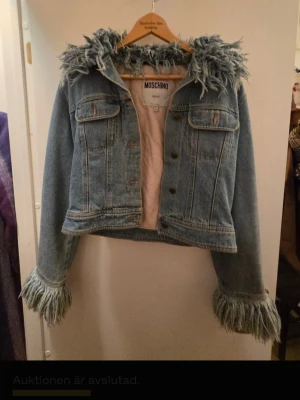 Urtuff Blå jeansjacka från Moschino - Unik blå jeansjacka från Moschino med ljusrosa foder. Jackan har dekorativ fuskpäls runt huvan och vid ärmsluten, samt två bröstfickor med lock. Croppad modell med coola detaljer som sticker ut.