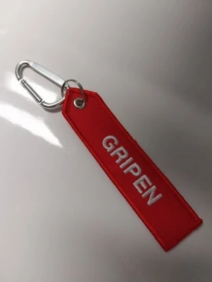 Saab Gripen Tagg med karbinhake - Säljer en röd tygnyckelring med vit broderad text 'GRIPEN' på ena sidan och 'REMOVE BEFORE FLIGHT' på andra sidan. Nyckelringen har en metallkarbin och ring, perfekt att hänga på väskan eller nycklarna för en cool flygstol.  Saab gripen Tagg med karbinhake Text saab.com på haken Text Remove before flight på ena sidan 