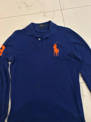 Blå långärmad pikétröja Polo Ralph Lauren - Blå långärmad pikétröja från Polo Ralph Lauren med stor orange broderad logga på bröstet och orange siffra på ärmen. Klassisk krage med knappar.