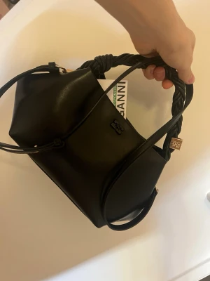 Svart handväska med flätat handtag Ganni - Snygg svart handväska från Ganni i slätt läder med ett unikt flätat handtag och guldfärgade detaljer. Väskan har en stilren design med diskret logga på framsidan och justerbar axelrem. Perfekt accessoar för att lyfta din outfit. (Jag accepterar budgivningar men var rättvis är ni snälla🙏🏼)