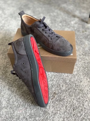 Mörkgråa Louboutin - Snygga mörkgråa sneakers i mocka från Christian Louboutin med ikonisk röd sula och svart snörning. Skorna har en låg profil och klassisk siluett som passar till många olika stilar. Perfekta för dig som vill sticka ut med exklusiva detaljer.