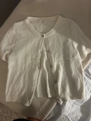 Vit kortärmad kofta från H&M - Söt vit kofta från H&M med korta ärmar och rund halsringning. Koftan har ribbade muddar och knäpps med små knappar framtill. Perfekt att slänga över en topp för en enkel och clean look.