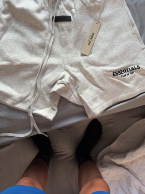 Gråa Essentials Fear of God shorts - Säljer ett par ljusgrå shorts från Essentials Fear of God med svart logga på benet. Shortsen har snörning i midjan och är tillverkade i mjuk bomull. Perfekta för en avslappnad och trendig stil.
